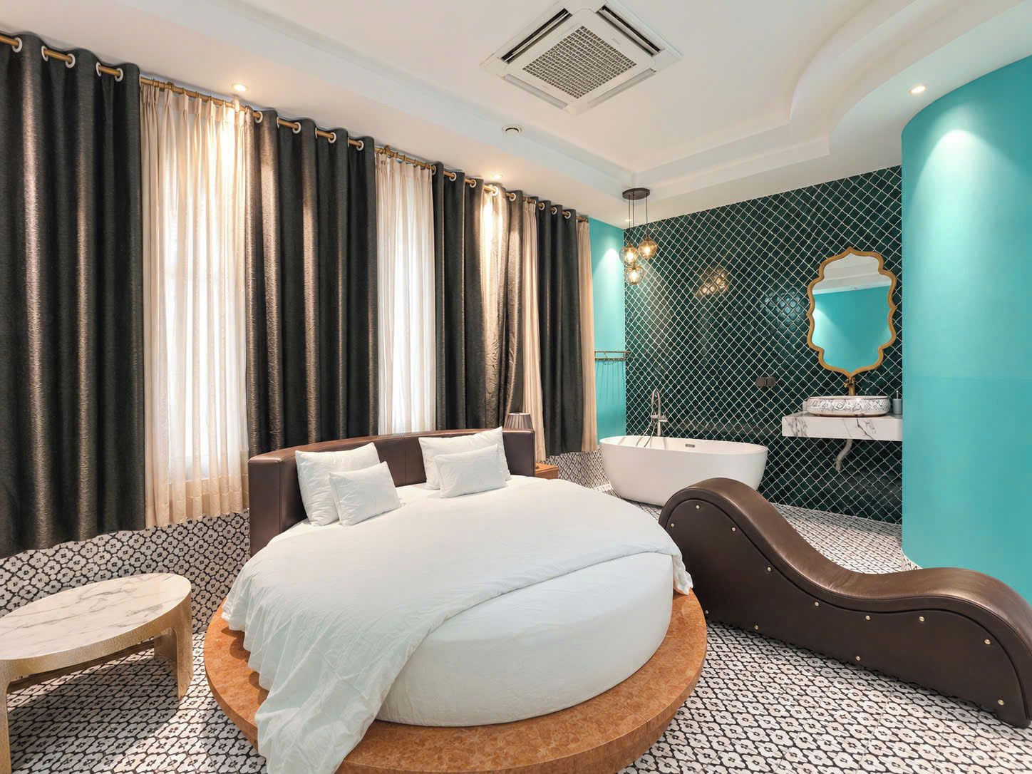 Una habitación de hotel moderna con una cama grande, una plataforma redonda de madera, una zona de estar, un baño con bañera independiente y un espejo decorativo.