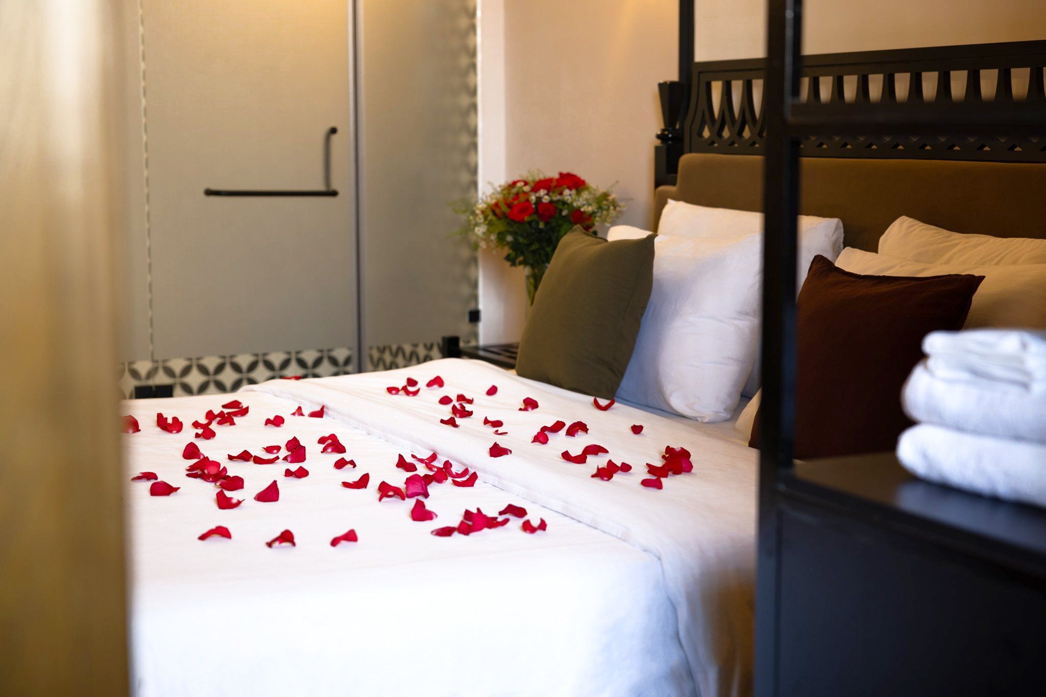 A hotel room with a bed, pillows, a nightstand, and a vase with flowers.