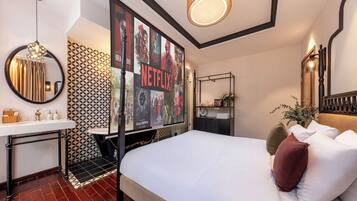 Ein Hotelzimmer mit einem großen Bett, einem Nachttisch mit Lampe, einem Spiegel, einem an der Wand befestigten Fernseher, der Filmplakate zeigt, und einer Wand mit Muster.