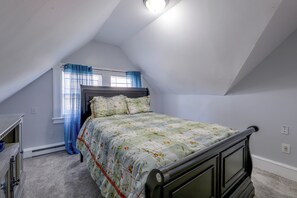 A bedroom with a sloped ceiling, a bed with floral bedding, a dresser, and a window with blue curtains. - Extended Springfield Stay Near Downtown & MGM (Springfield)
