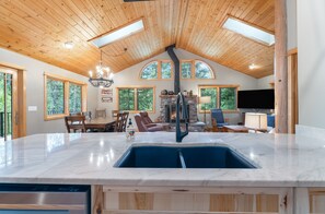 A spacious kitchen with a large island, a stone fireplace, and a dining area with wooden furniture. - NEW | 3 Mi to Whitefish Mountain | Ski + Snowshoe | Cedar Home, Stone Fireplace, Sleeps 8 (Whitefish)