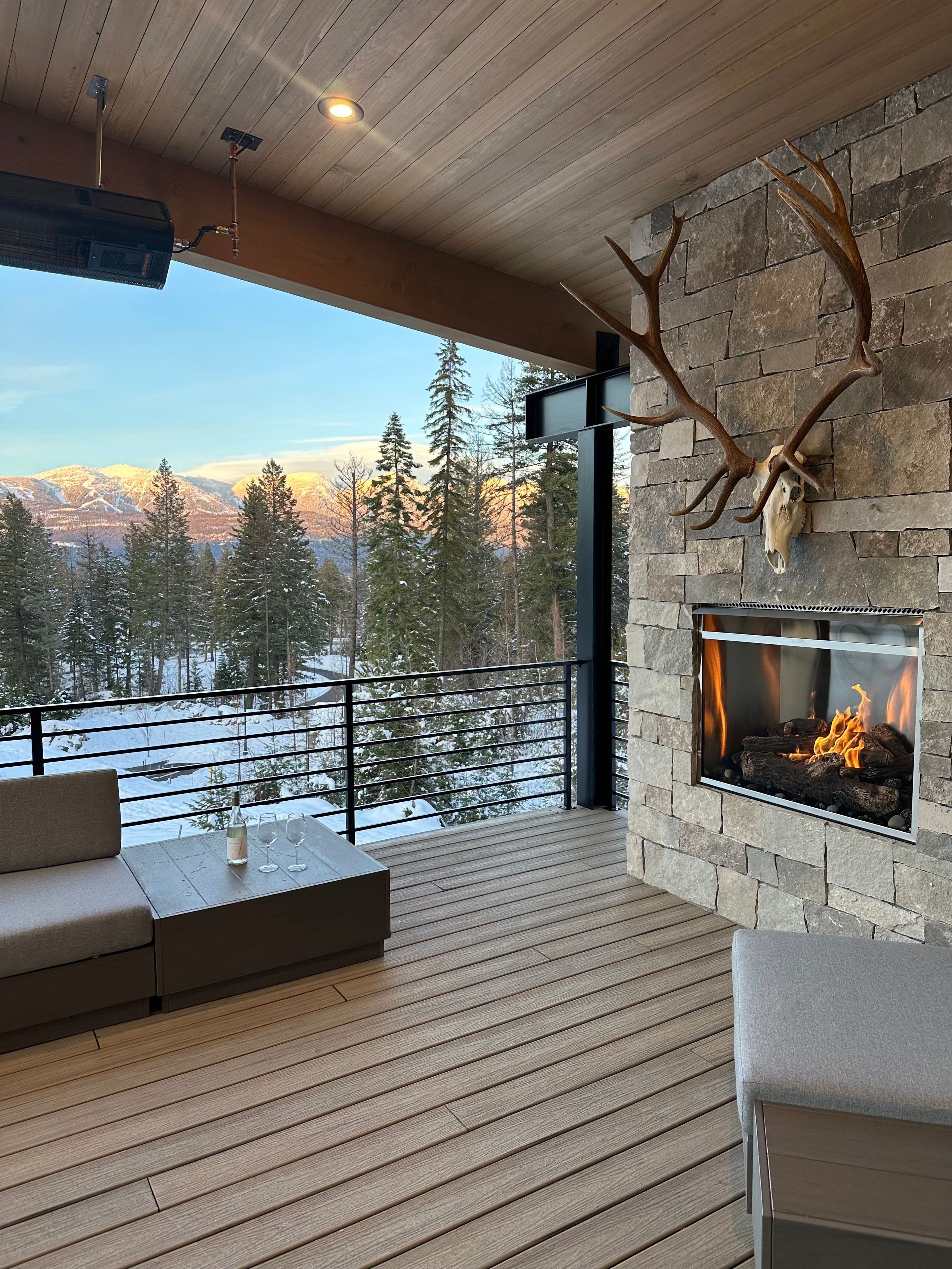 A deck with a stone fireplace, a mounted deer skull, and a view of snow-covered mountains.