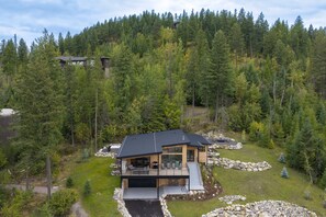 A modern cabin with a large deck, surrounded by dense forest. - New! Family Ski Getaway with Hot Tub, Chef's Kitchen & Fireside Deck, Monthly Stays (Whitefish)