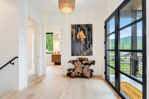 A modern interior with a wooden floor, a unique console table, and a large window offering a view of a forest. - New! Family Ski Getaway with Hot Tub, Chef's Kitchen & Fireside Deck, Monthly Stays (Whitefish)