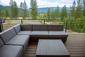 A sectional sofa and a small table on a wooden deck with a view of a forest and mountains. - New! Family Ski Getaway with Hot Tub, Chef's Kitchen & Fireside Deck, Monthly Stays (Whitefish)