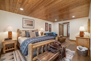 A rustic bedroom with a wooden ceiling, a bed with a blue blanket, two side tables with lamps, and a door leading to another room. - New Listing! Ski-In/Ski-Out with Indoor Hot Tub in Big Sky (Big Sky)
