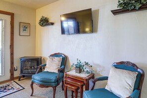 Un salon avec deux fauteuils bleus, une petite table en bois sur laquelle est posé un vase, un téléviseur à écran plat fixé au mur et un poêle à bois ancien.