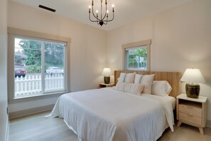 Une chambre à coucher avec un grand lit, deux tables de chevet avec des lampes, un lustre et deux fenêtres donnant sur l’extérieur.