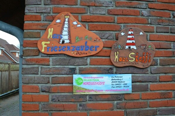 Dos letreros decorativos en una pared de ladrillo, uno con la etiqueta 'Fiesenzauber' y el otro con 'Hans Stett'. Debajo de los letreros, hay una tarjeta de presentación con la información de contacto para 'Vermögungsservice' y 'NORDSEEKÜSTE'.