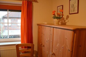A room with a wooden cabinet, a chair, a window with orange curtains, and a vase with flowers on the cabinet. - Holiday home in Hooksiel with swimming pool (Hooksiel)