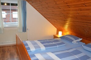 A bedroom with a sloped ceiling, a bed with blue and white bedding, a nightstand with a lamp, and a window with curtains. - Holiday home in Hooksiel with swimming pool (Hooksiel)