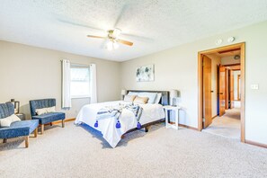 Un dormitorio con cama, dos sillas, ventilador de techo, ventana con cortinas, mesita de noche y puerta que da a otra habitación.
