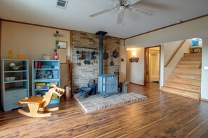 A room with a brick wall, a wooden floor, a ceiling fan, a bookshelf, a rocking horse, and a fireplace with hanging pots. - 3 Mi to Norfork Lake! Multi-Level Haven for Groups (Mountain Home)