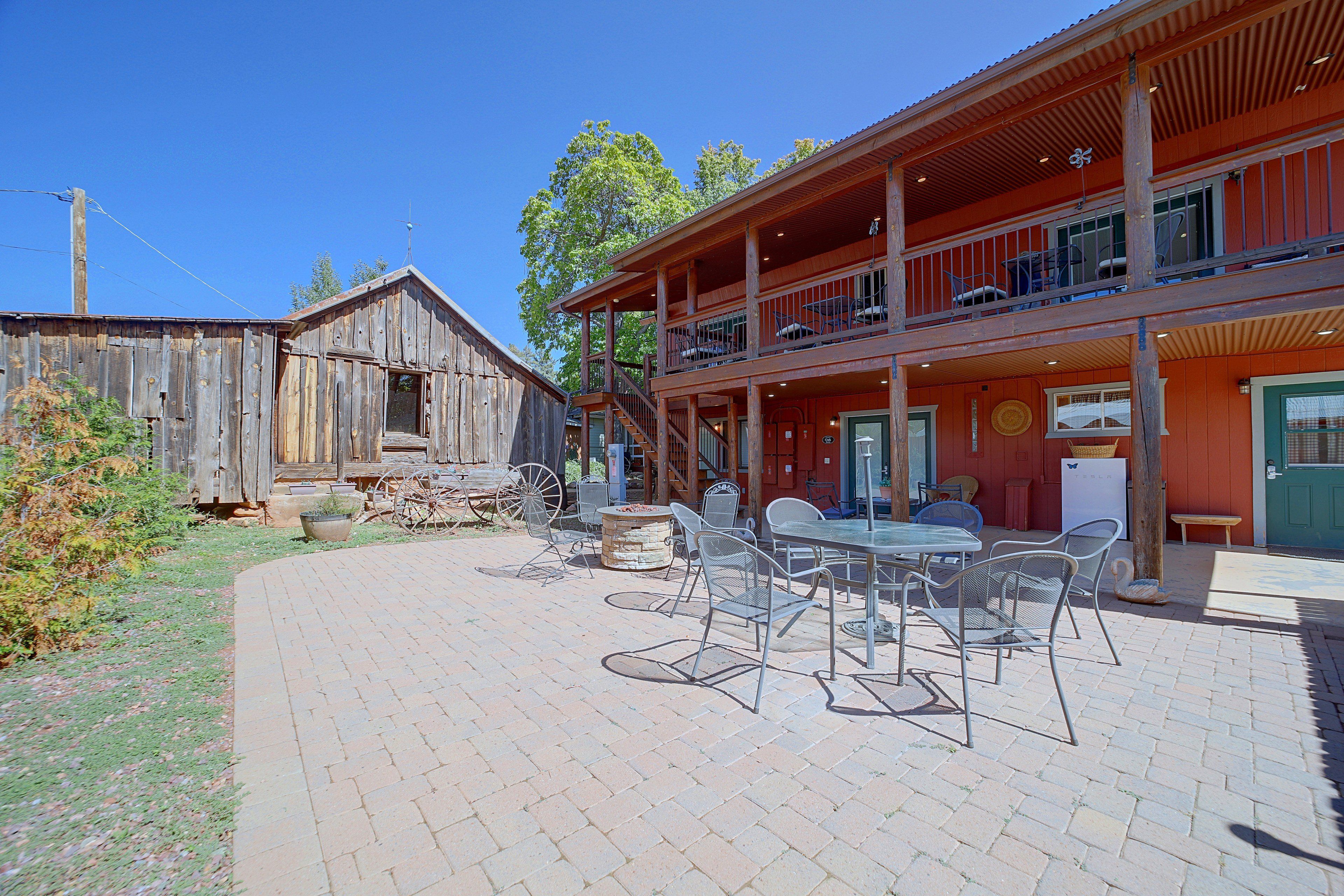 Uma área de estar rústica ao ar livre, com cadeiras de metal e uma mesa, um edifício de madeira com varanda e um prédio vermelho com porta verde.