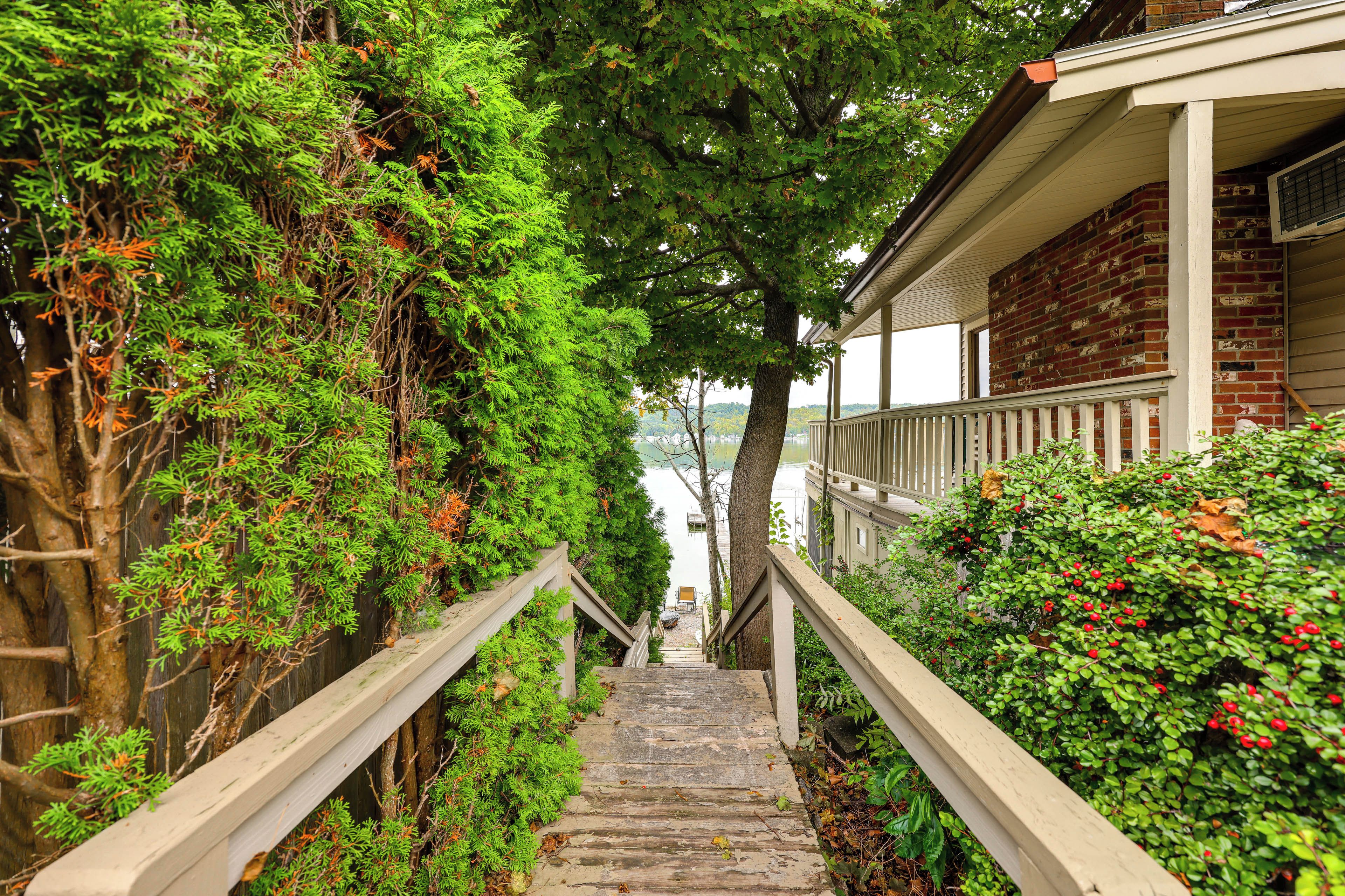 A wooden staircase leading to a building with a brick facade, surrounded by lush greenery and a view of a body of water.