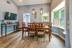 A dining area with a wooden table and chairs, a television mounted on the wall, and a window with a view of greenery. - Steps to Lake! Home w/ Deck & Views in Conesus (Conesus)