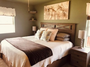 Un dormitorio con cama, lámparas de noche, mesita de noche, una obra de arte en la pared y una ventana con cortina.