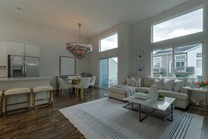 Een ruime woonkamer met een groot raam, een kroonluchter en een moderne keuken met een koelkast.