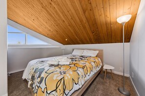 A bedroom with a sloped ceiling, a bed with a floral bedspread, a bedside table, and a floor lamp. - 489 Tahoe Keys Blvd, Unit 69 1 Bedroom Condo (South Lake Tahoe)
