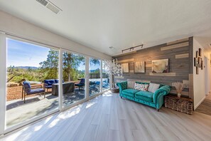 Ein geräumiger Wohnbereich mit einer großen Schiebeglastür, die auf eine Terrasse im Freien führt, einem türkisfarbenen Sofa und einer Holzwand mit Kunstwerken.