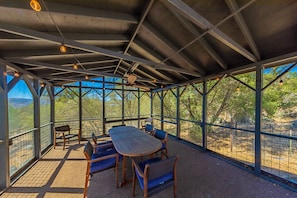 Ein überdachter Außenbereich mit Tisch und Stühlen, umgeben von einem Metallzaun, mit Blick auf eine natürliche Landschaft.