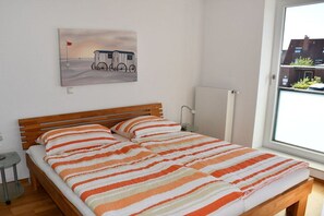 A bedroom with a wooden bed, striped bedding, a small bedside table, a radiator, and a window with a view of buildings. - Holiday apartment with 1 bedroom (Norderney)