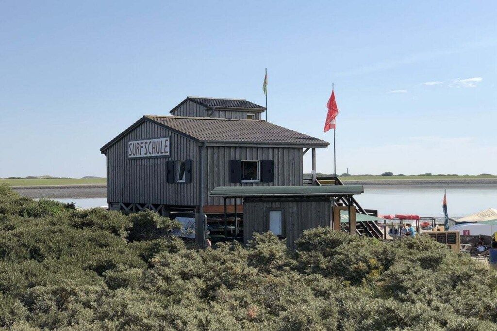 Un bâtiment en bois avec une enseigne indiquant « SURFSCHULE » et un mât sur lequel flotte un drapeau.