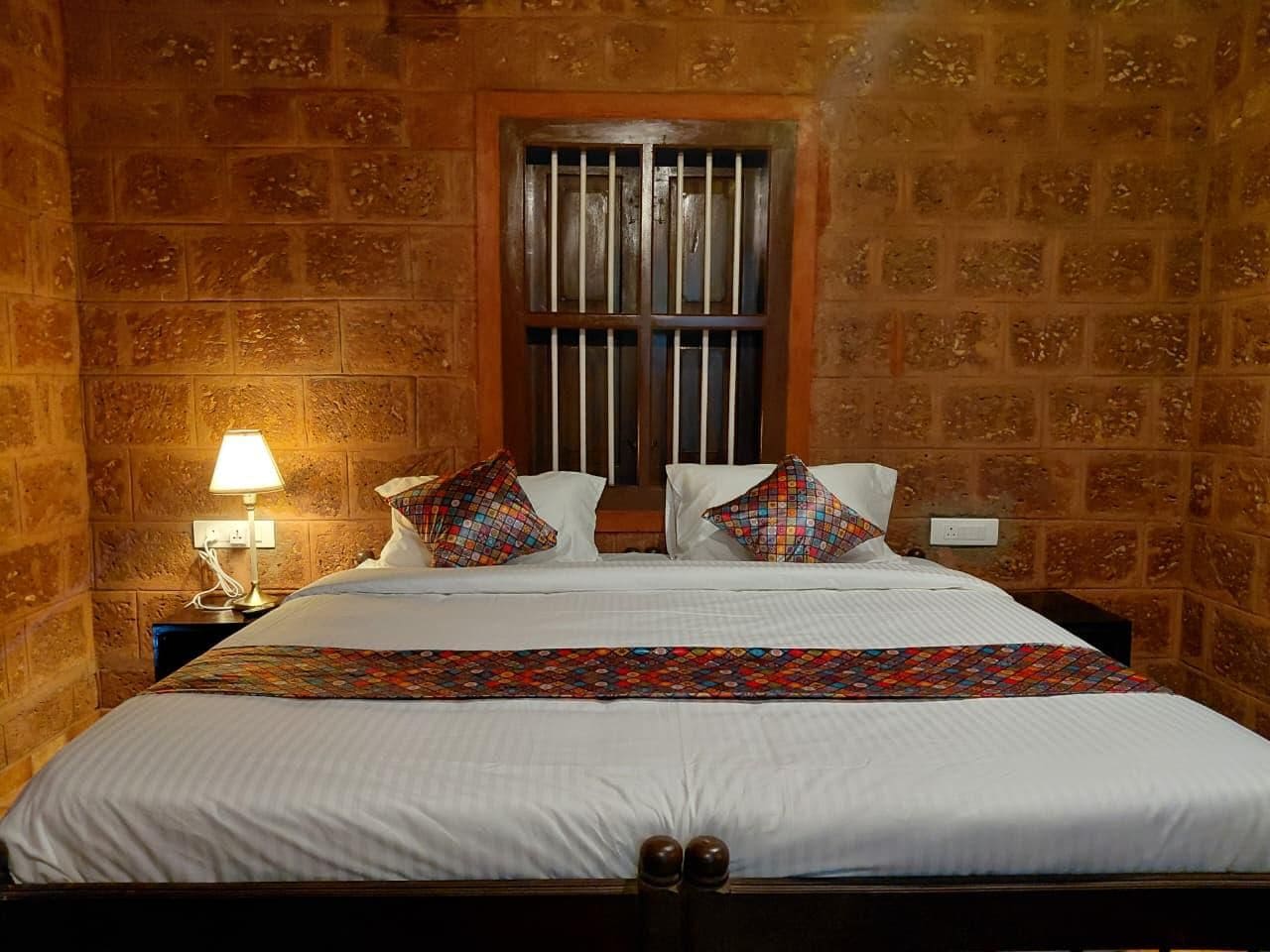 A hotel room with a brick wall, a window with bars, a bed with white linens, two pillows, a bedside lamp, and a patterned throw.