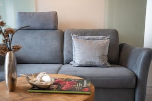 Ein graues Sofa mit einem Kissen, ein kleiner Tisch mit einem Buch und einer dekorativen Schale, und eine Vase mit getrockneten Blumen.