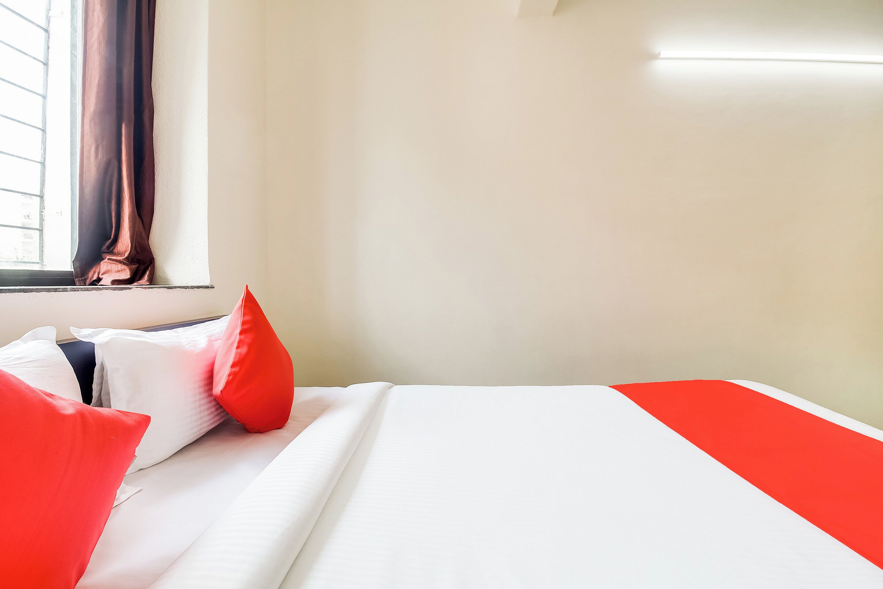 A hotel room with a bed, pillows, a window with curtains, and a wall-mounted light.