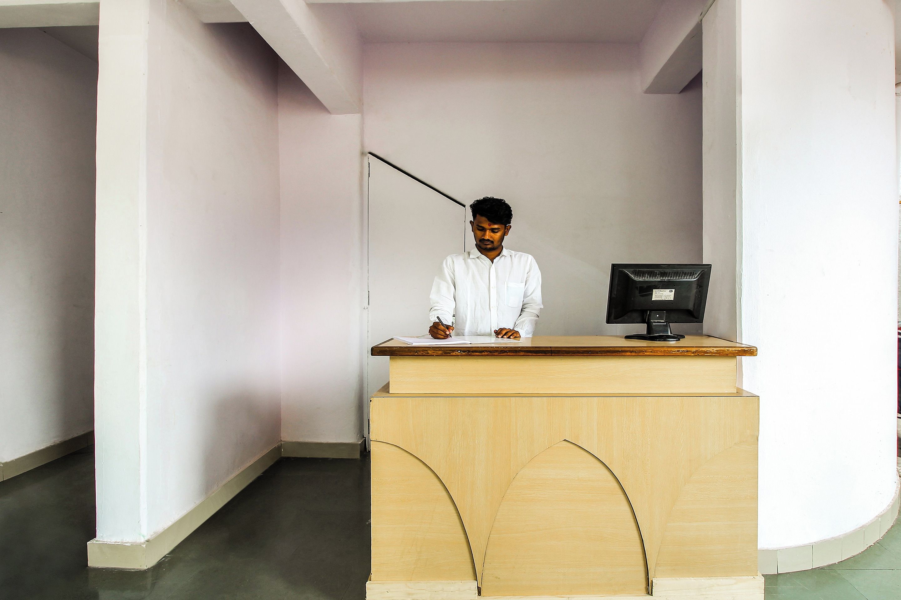 A person seated at a wooden desk with a computer monitor, in a room with white walls and a tiled floor.