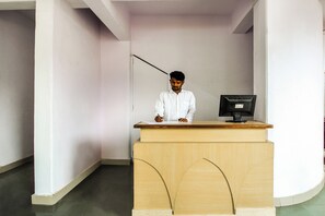 A person seated at a wooden desk with a computer monitor, in a room with white walls and a tiled floor. - Hotel O Galaxy Near INOX (Pune)