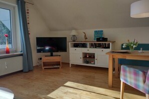 A living room with a television, wooden furniture, a shelf with books, and a window with curtains. - Holiday apartment with 1 bedroom (Büsumer Deichhausen)