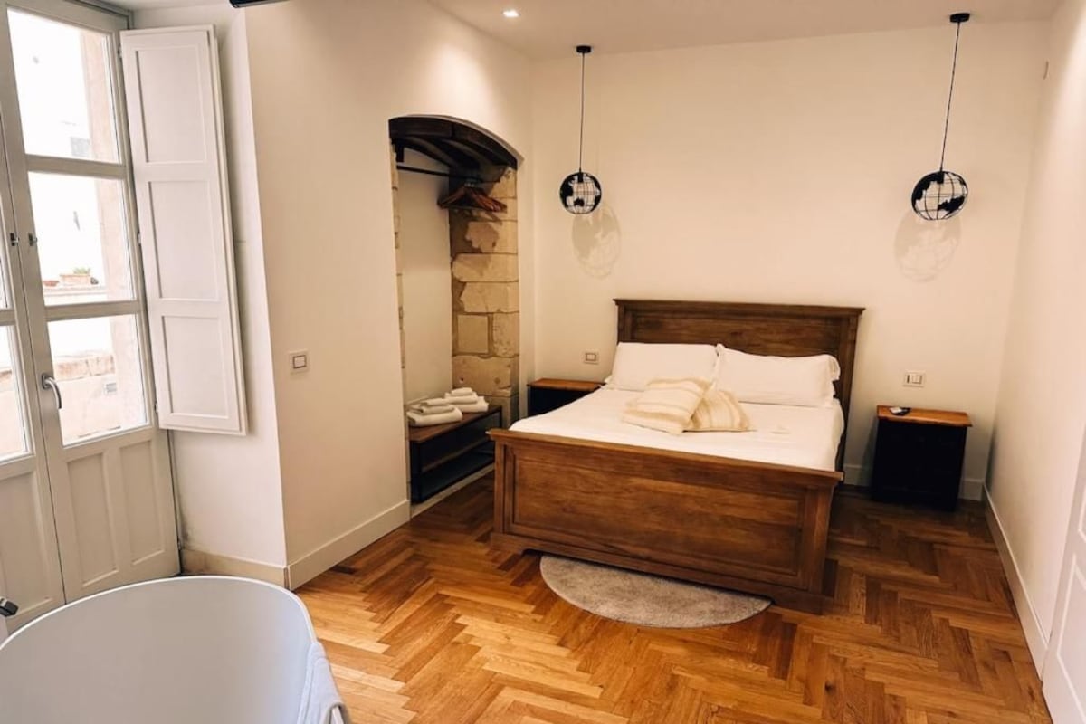 Quarto com cama de madeira, parede de pedra, banheira branca e janela com vista.