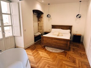 Quarto com cama de madeira, parede de pedra, banheira branca e janela com vista.
