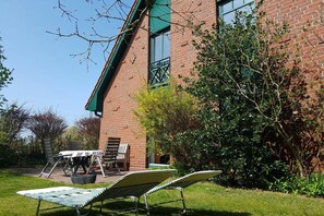 A brick building with a green roof, a patio with a table and chairs, and a garden area with a lounge chair. - Holiday Residence Oystercatcher II, 1a (Spiekeroog)