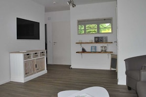 A room with a television mounted on the wall, a wooden cabinet with drawers, a window with a view, and a grey sofa. - Holiday apartment with 2 bedrooms (Norddeich)