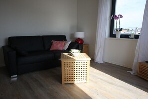 A living room with a black sofa, a wooden side table, a red lamp, and a window with curtains. - Holiday apartment in Husum with swimming pool (Husum)