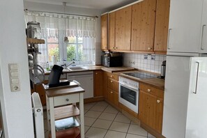 A compact kitchen with wooden cabinets, a built-in oven, a sink, and a window with curtains. - Holiday home with 4 bedrooms (Carolinensiel)