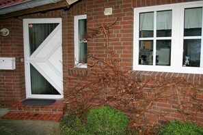 Un bâtiment en briques avec une porte et des fenêtres blanches, un petit jardin et un chemin en briques menant à l’entrée.