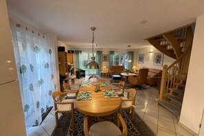 A dining area with a wooden table, chairs, and a chandelier. There is a staircase to the right and a living area with a sofa and lamp. - Holiday home with 4 bedrooms (Carolinensiel)
