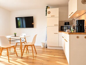 A modern kitchen with white cabinetry, a flat-screen TV mounted on the wall, and a dining area with a table and chairs. - Modern Apartment with Stunning Mountain Views @ Sunrise Pfronten (Pfronten)