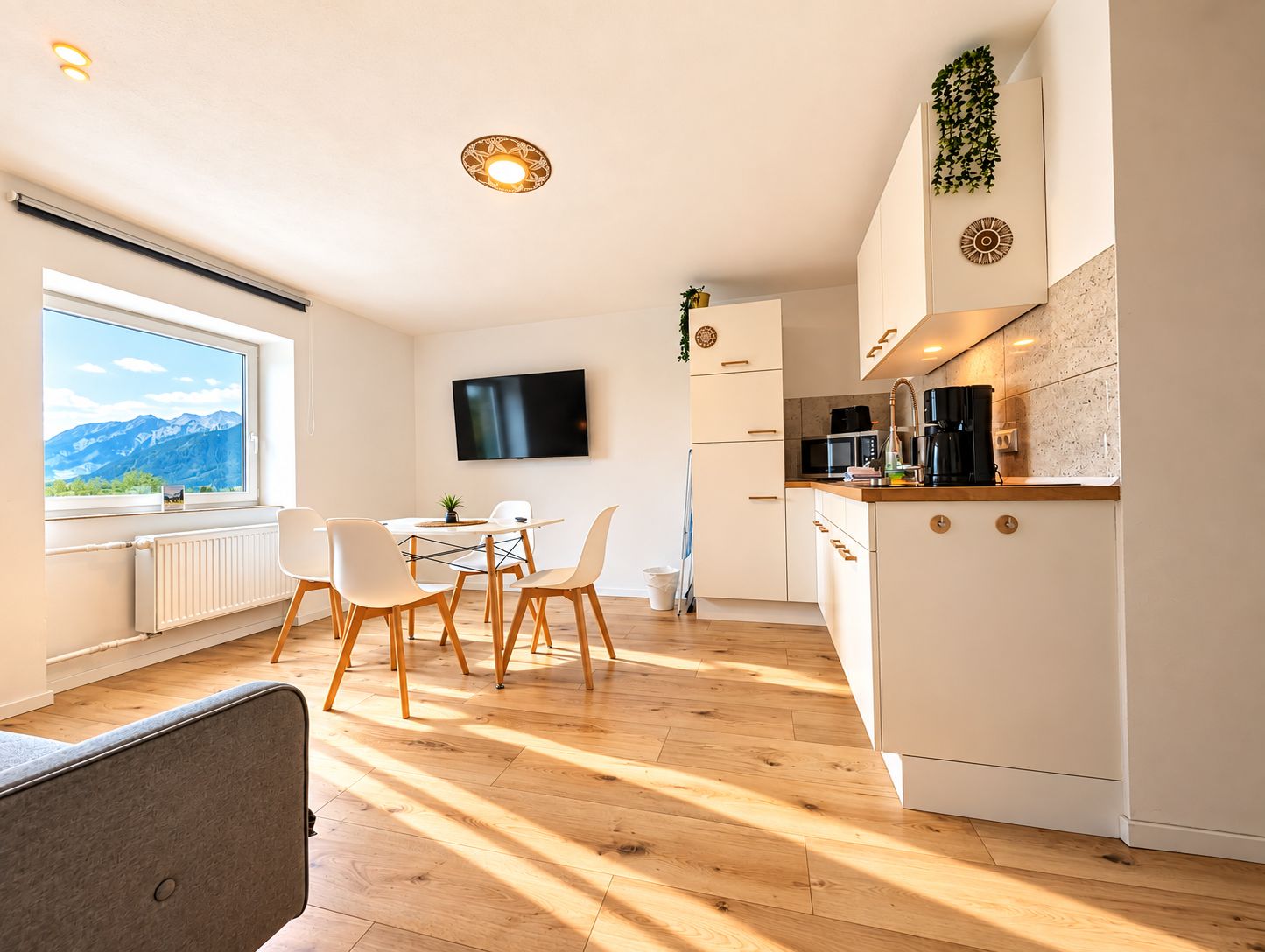 A modern kitchen with a dining area, a TV mounted on the wall, and a view of mountains through the window.