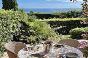 Un espace de restauration en plein air avec une table dressée pour quatre personnes, entouré d’une végétation luxuriante et offrant une vue sur la mer.
