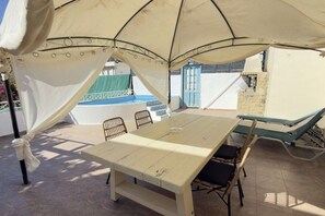 A covered outdoor dining area with a table and chairs, a reclining chair, and a view of a pool. - Villa in Pitsidia with swimming pool (Pitsidia)