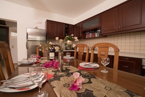 Ein Essbereich mit einem langen Tisch, der für eine Mahlzeit gedeckt ist, Holzstühlen, einem Kühlschrank und einem Schrank mit Büchern.