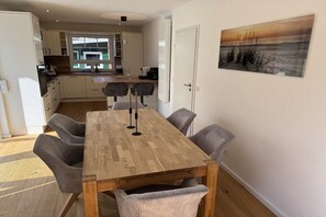 A dining area with a wooden table and cushioned chairs, a kitchen in the background, and a painting on the wall. - Holiday HOME "North Sea" (Hooksiel)
