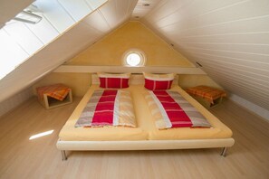 A room with a sloped ceiling, a bed with two pillows, a small round window, and a wooden floor. - Atlantic beach apartment (Norderney)