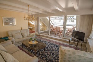 A living room with a sofa, coffee table, and a TV on a stand. There is a balcony with chairs and a table. - Atlantic beach apartment (Norderney)
