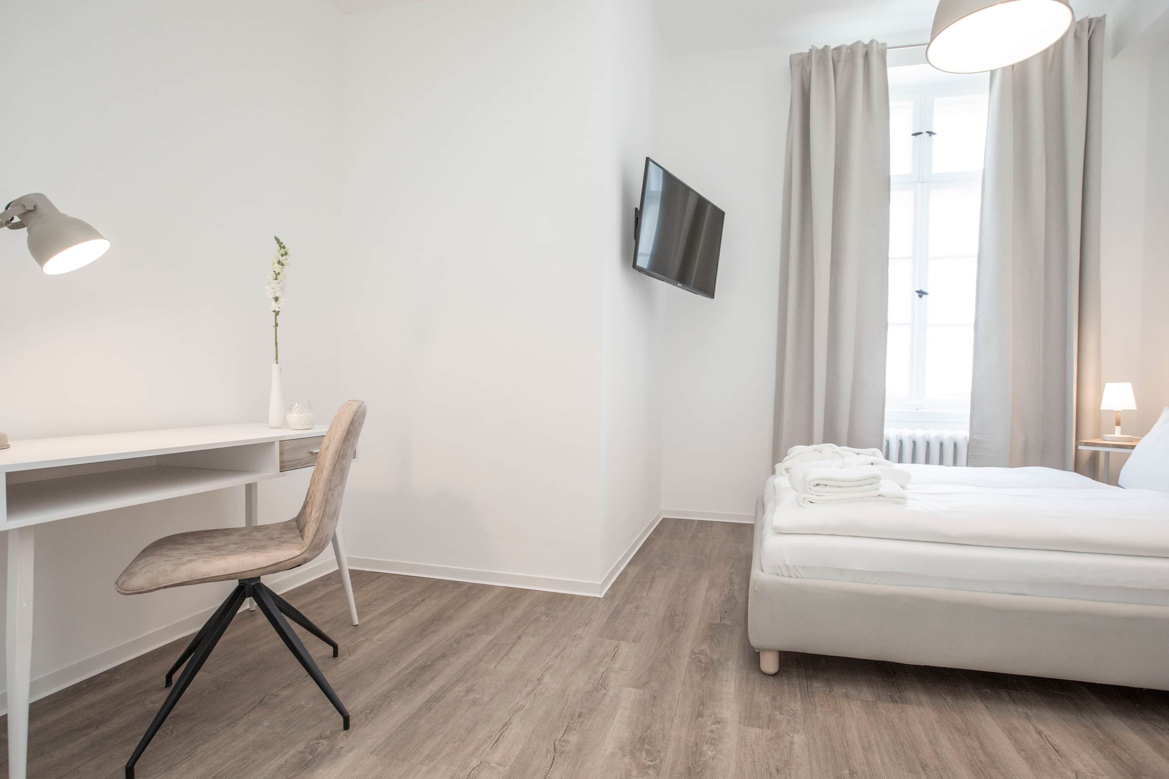 Ein Hotelzimmer mit einem Bett, einem Schreibtisch mit Stuhl, einem an der Wand montierten Fernseher und einem Fenster mit Vorhängen.